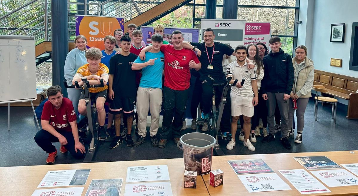 a group of serc students and employees grouped together in a row, two sitting a spin bike, a table with a fundraising bucket in the foreground